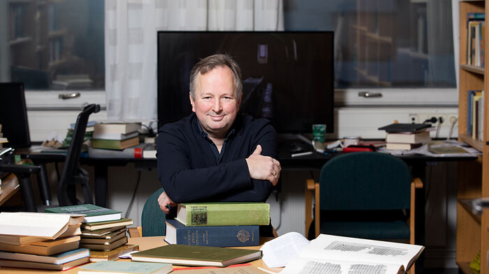 Foto av Hugo Lundhaug som sitter ved et bord på sitt kontor og lener seg på en stabel med bøker